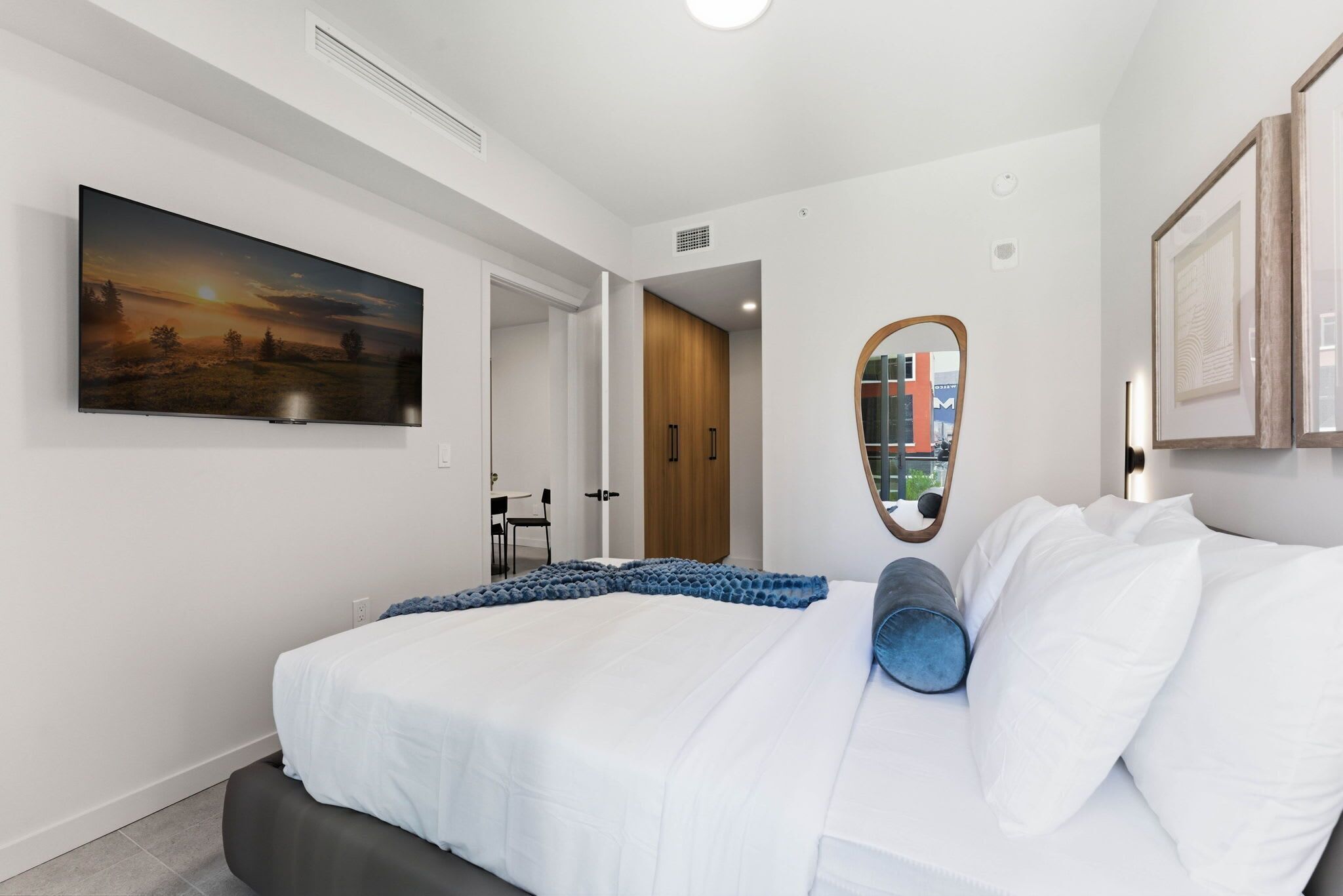 A modern bedroom with a large bed, a painting, a mirror, and a view of the city through the window.