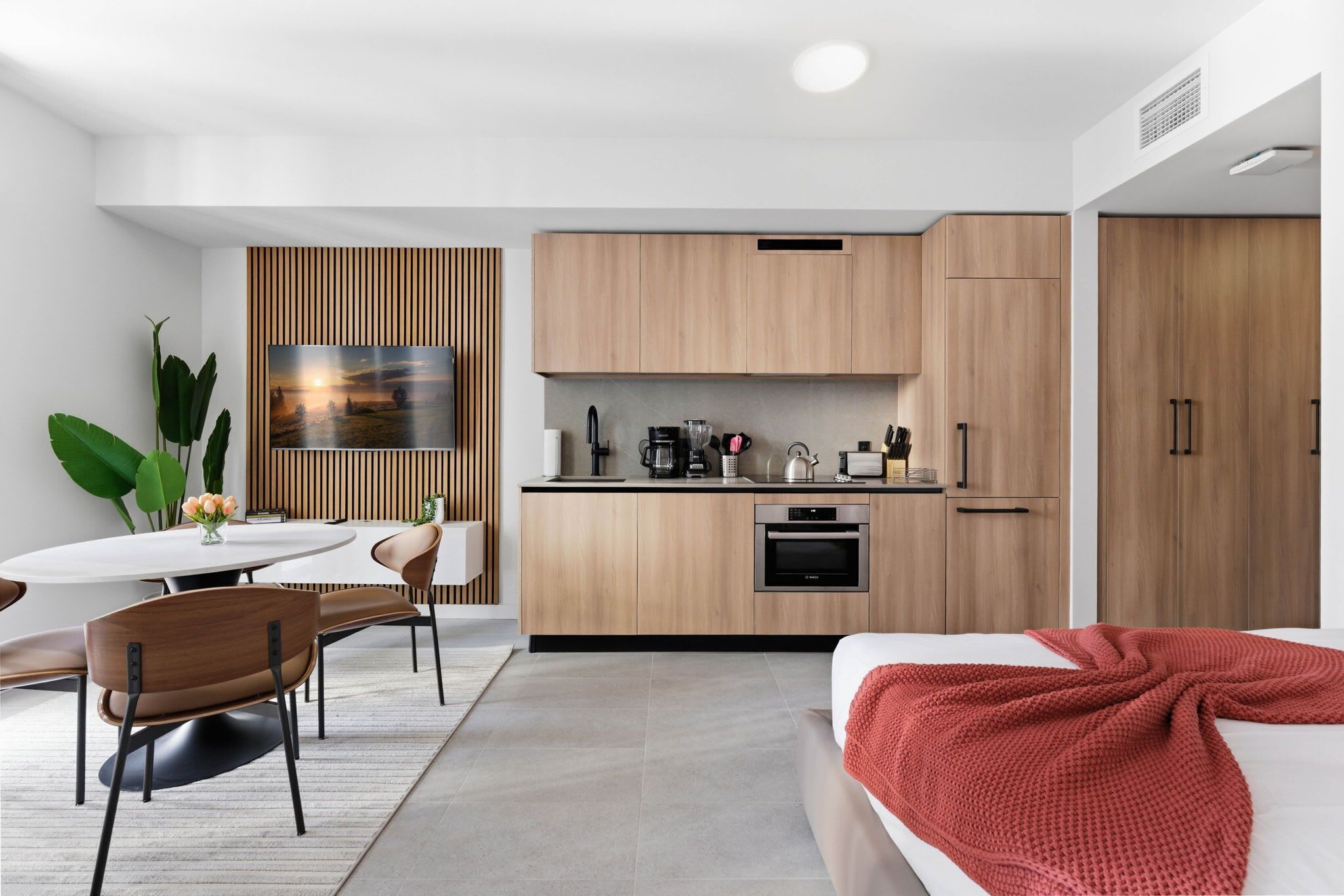 Ein modernes, kompaktes Hotelzimmer mit Küchenzeile, Essbereich und einem Bett mit roter Decke.