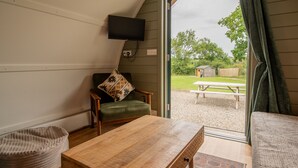 A room with a wooden table, a green chair, a beige bean bag, a television mounted on the wall, and a view of an outdoor picnic area. - Luxury Cabin with Hot Tub and Pergola in Yorkshire (Northallerton)