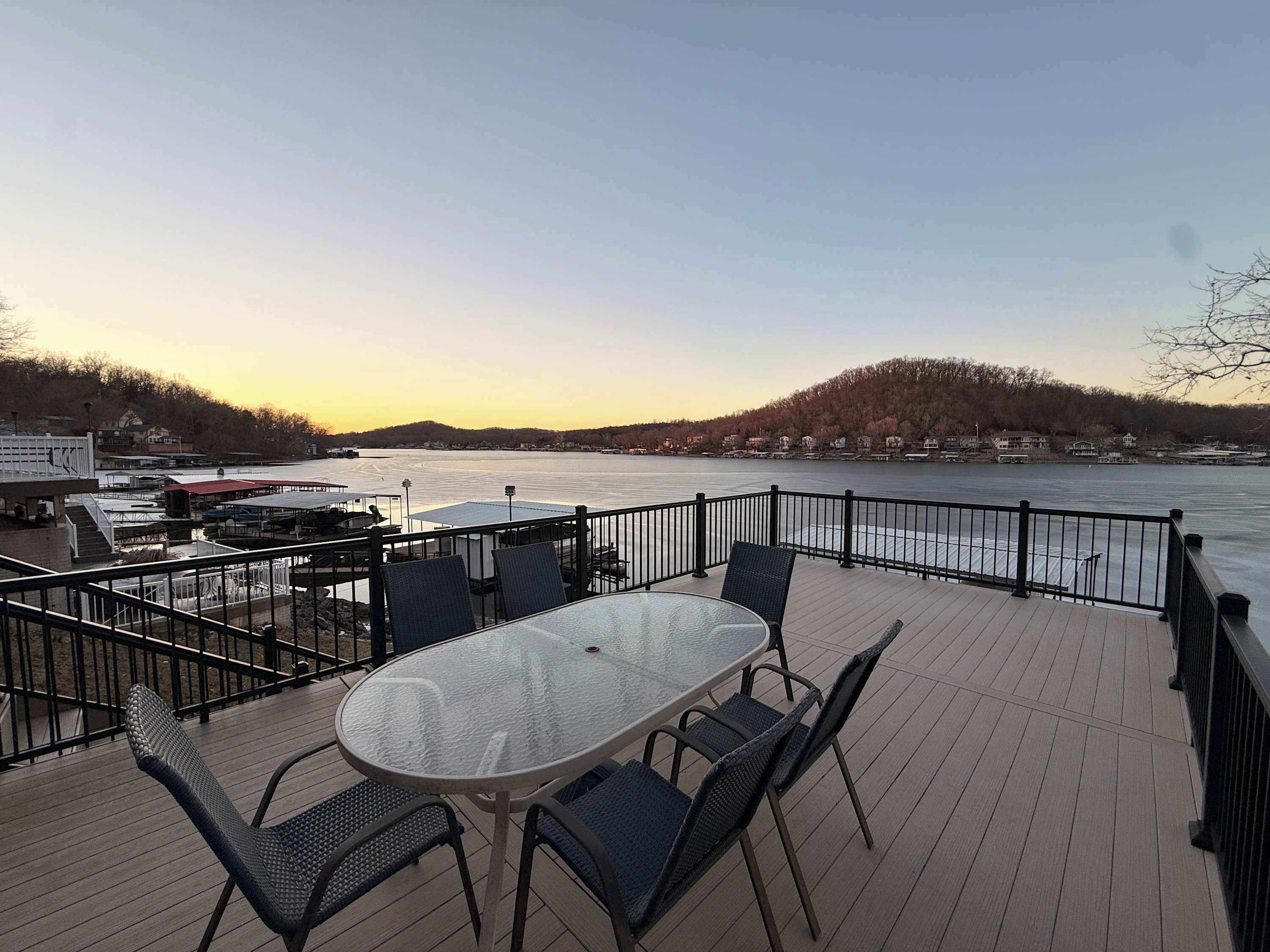 A deck with a glass table and chairs overlooking a river, with a view of houses and trees in the background.
