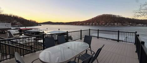 A deck with a glass table and chairs overlooking a river, with a view of houses and trees in the background.