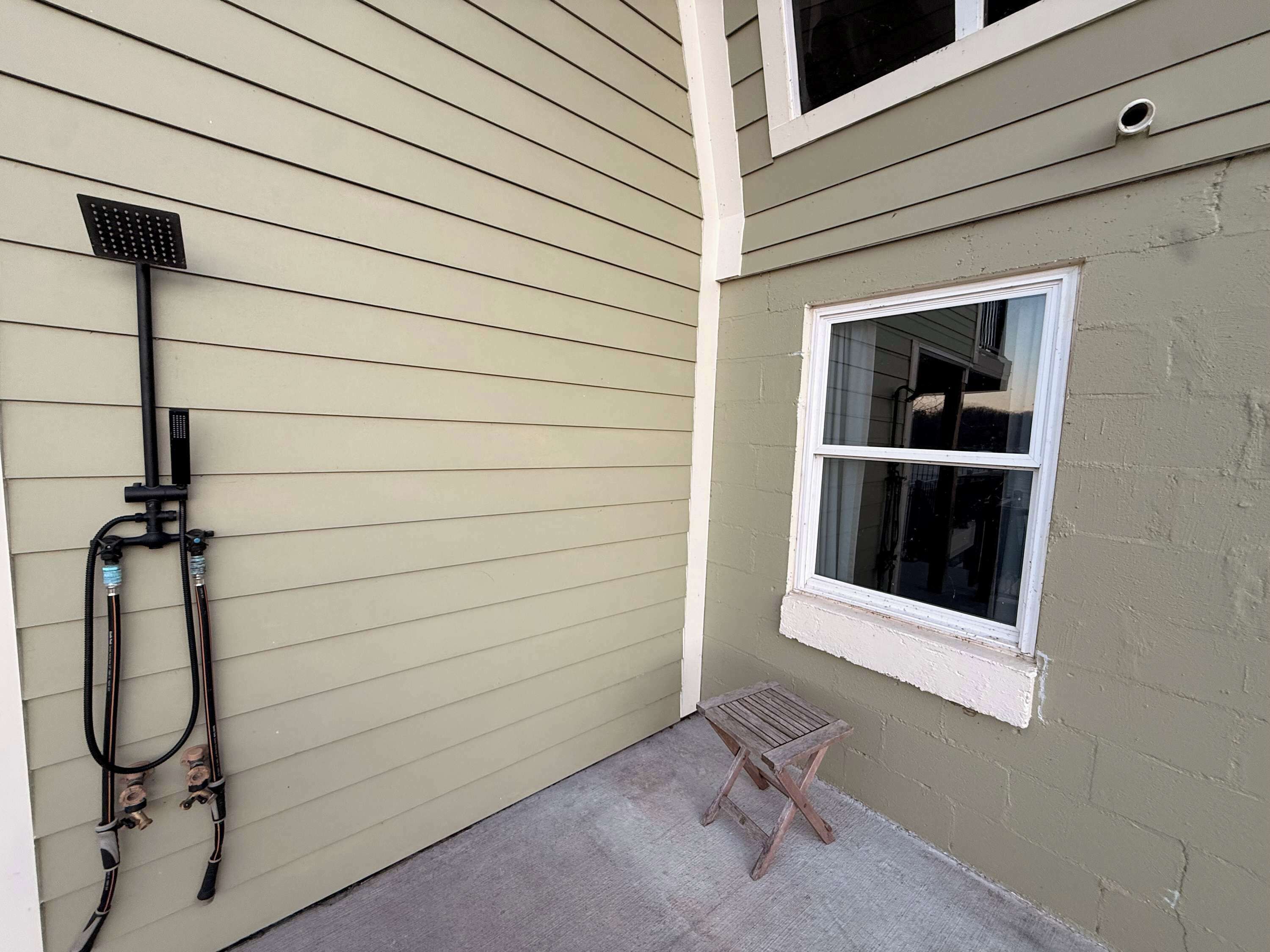 A small outdoor area with a window, a wooden stool, and a hose attached to a wall.