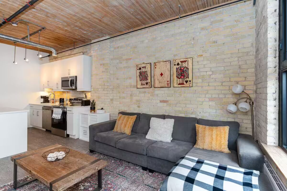 Un salon chaleureux avec un canapé gris, une table basse en bois et un tapis à motifs. On aperçoit, au fond, une cuisine avec des meubles blancs et un four à micro-ondes.