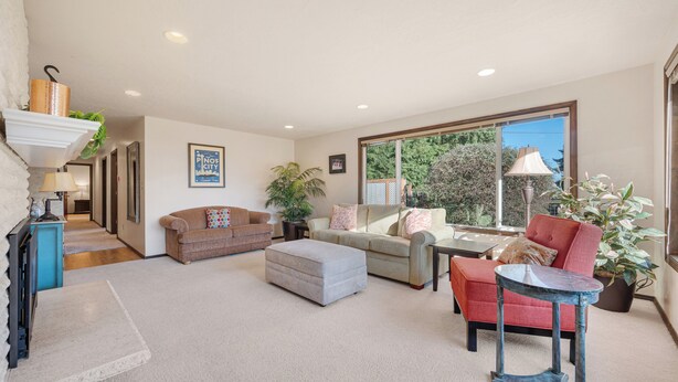A spacious living room with a large window, a fireplace, and a variety of seating options including a red chair, a grey ottoman, and a beige sofa. - Oregon Wine Country Hillside Retreat w/4 bedrooms, 3 baths. (Dundee)