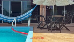 A pool area with a blue and white hammock, a red floating pool toy, and a wooden table with chairs. - Apto Arraial do Cabo c/ Piscina e Bilhar (Arraial do Cabo)