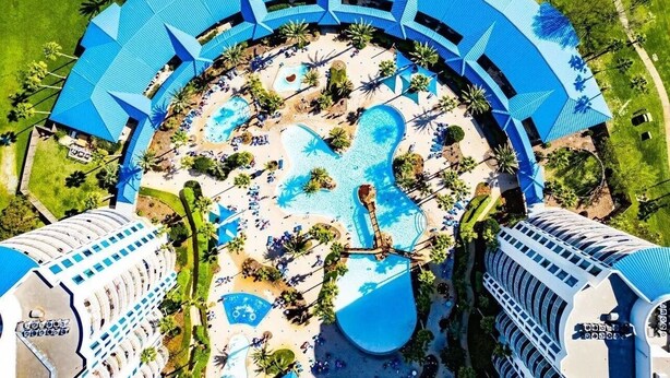 Vue aérienne d’un complexe hôtelier avec une piscine centrale entourée de palmiers et de chaises longues.