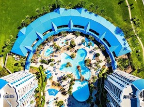 Luftaufnahme eines Resorts mit einem zentralen Pool, umgeben von Palmen und Liegestühlen.