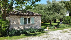 Una piccola casa in pietra con tetto in tegole, immersa in un giardino curato con diverse piante e un vialetto sterrato.