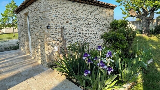 Un edificio in pietra con tetto in tegole, circondato da un giardino con fiori viola e piante verdi.