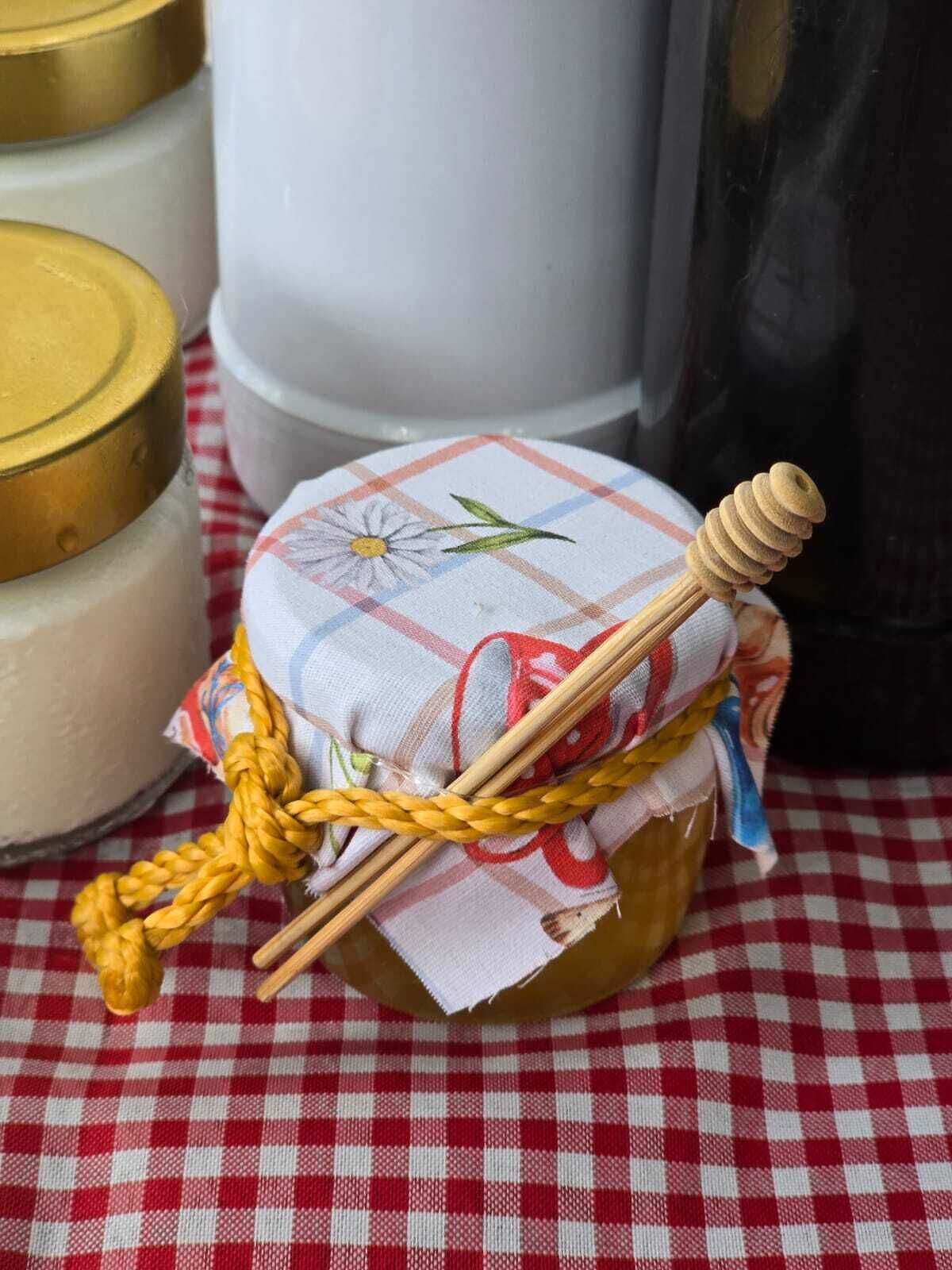Um frasco de mel com um regador de mel e um pano amarrado ao redor, colocado sobre uma toalha de mesa xadrez.