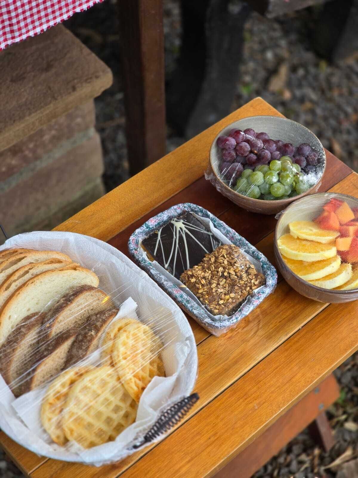 Et træbord med ternet dug, der indeholder en kurv med skiveskåret brød, en krukke med granola, en skål blandede druer og en skål med frugtstykker.