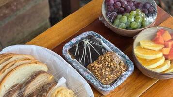 Et træbord med ternet dug, der indeholder en kurv med skiveskåret brød, en krukke med granola, en skål blandede druer og en skål med frugtstykker.