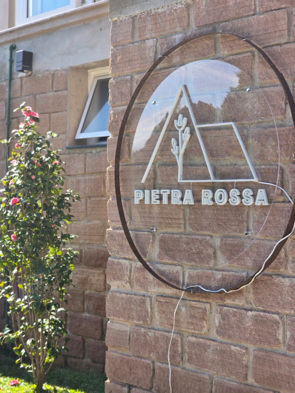 Una señal con el nombre 'PIETRA ROSSA' en una pared de ladrillo, junto a una pequeña planta en maceta.