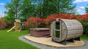 Una vasca idromassaggio in legno su un piedistallo, una casetta in legno con uno scivolo e un giardino con arbusti dai colori autunnali.
