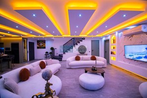 Un salon moderne avec des canapés blancs, un pouf rond et un téléviseur fixé au mur.