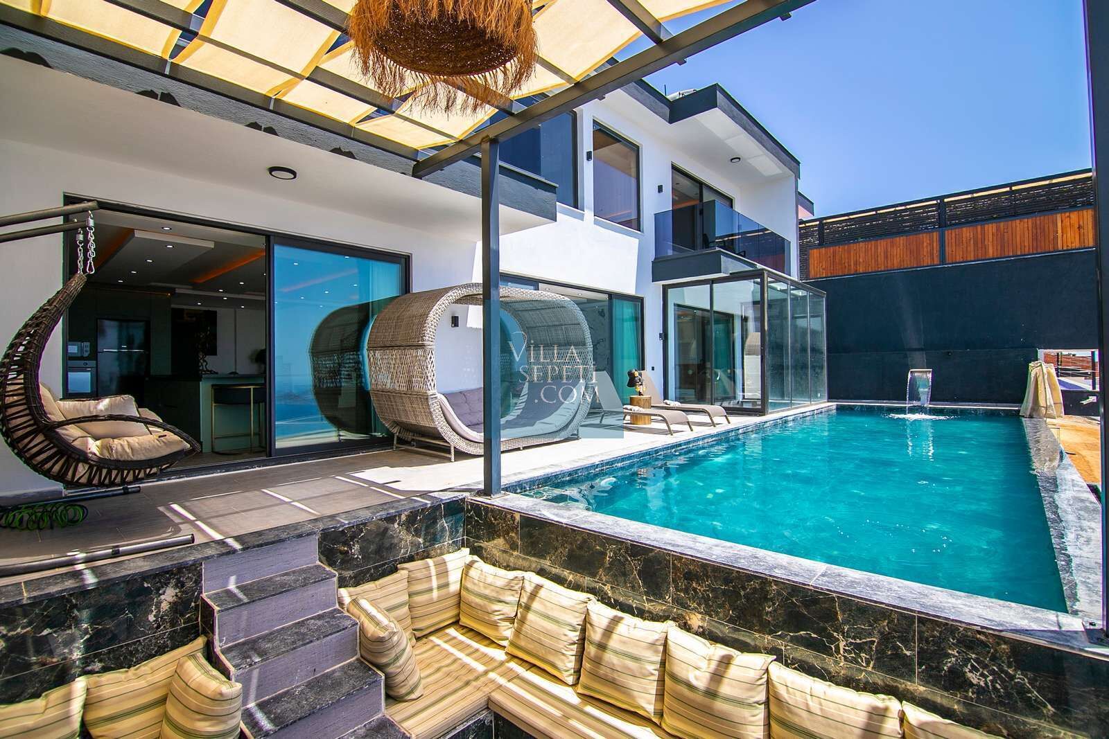 A modern outdoor pool area with a glass-enclosed lounge, a hanging chair, and a seating area with cushions.