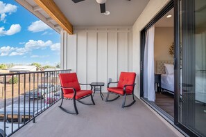 Un balcón con dos sillas rojas y una mesita, una cama con ropa de cama blanca, y vistas al cielo y edificios lejanos.