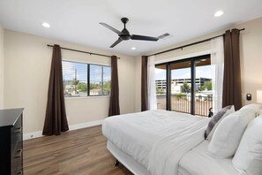 Un dormitorio con cama, ventilador de techo, ventana con cortinas y vistas a edificios.