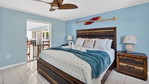 Quarto com cama, cabeceira de madeira, duas mesas de cabeceira, um ventilador de teto e vista para área externa por uma porta de vidro.