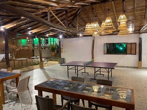 An indoor space with tables and chairs, a ping pong table, and hanging lamps.