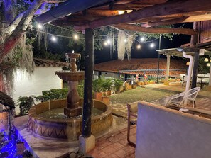 A nighttime outdoor area with a fountain, string lights, and seating under a wooden pergola.