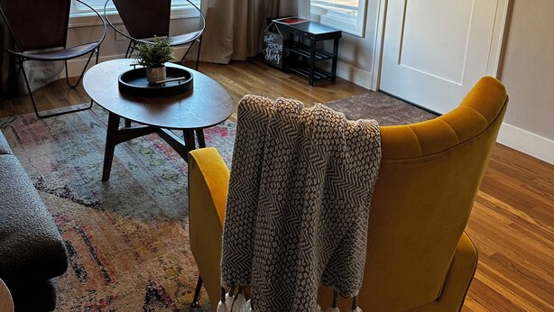 A living room with a yellow armchair, a round coffee table, and a sofa. There are two windows with curtains, a door, and a small table with a sign.