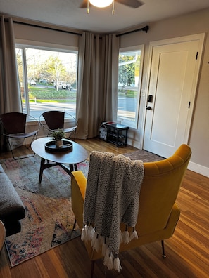 Een woonkamer met een gele fauteuil, een ronde salontafel en een bank. Twee ramen met gordijnen, een deur en een kleine tafel met een bordje.