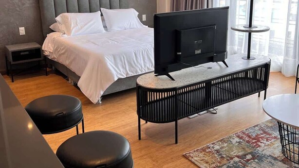 Una habitación de hotel moderna con una cama, un televisor, una consola negra, dos taburetes negros y una alfombra estampada.