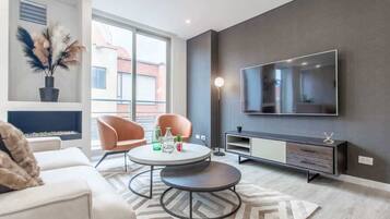 Ein stilvolles Wohnzimmer mit einer Couch, zwei Sesseln, einem runden Couchtisch, einem flachbildfernseher an der Wand, einer Anrichte und einer Topfpflanze.