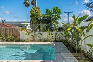 A swimming pool with a clear blue water surface, surrounded by various tropical plants and a wooden fence.