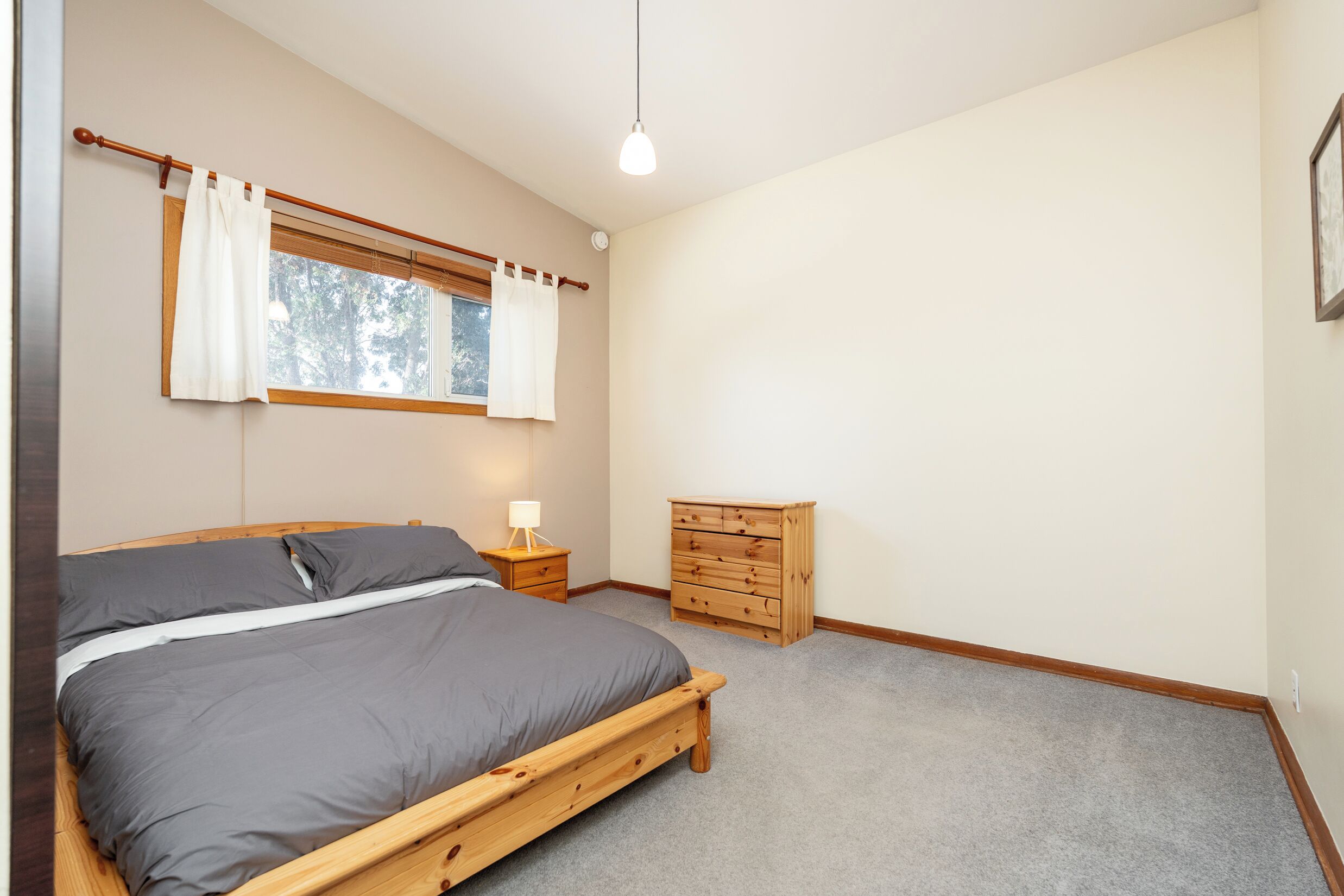 A bedroom with a wooden bed, a nightstand with a lamp, a dresser, and a window with curtains.