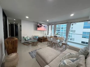 A modern living room with a sofa, dining table, chairs, a TV, and large windows offering a city view. - Charming 2BR Beachfront Apartment with AC in Morros, La Boquilla Cartagena. CTG (Cartagena de Indias)