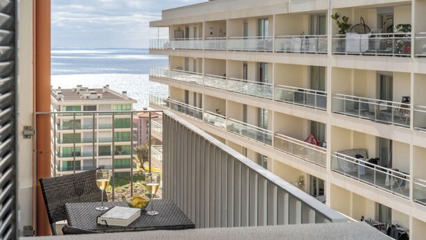 A balcony with a table, a glass of wine, and a book. The balcony overlooks a building with multiple balconies and a view of the sea. - Camélia's Apartment I –  Modern in Funchal (Funchal)