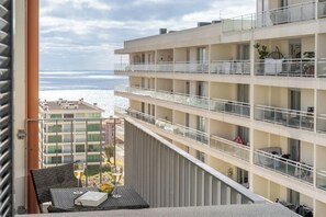 Un balcón con una mesa, una copa de vino y un libro. El balcón da a un edificio con varios balcones y vistas al mar.