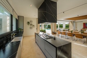 Eine moderne Küche mit einer zentralen Kochinsel, einem Essbereich mit Holztischen und Blick auf einen Pool.