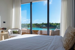 Ein Schlafzimmer mit einem großen Bett, Blick auf einen Pool und einem Korbsessel.