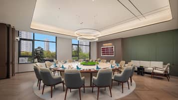 A modern conference room with a round table, chairs, and a view of buildings through large windows.