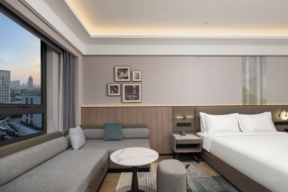 Una habitación de hotel moderna con un sofá, una mesita redonda pequeña, una mesita de noche con un reloj, una cama y vistas a la ciudad.