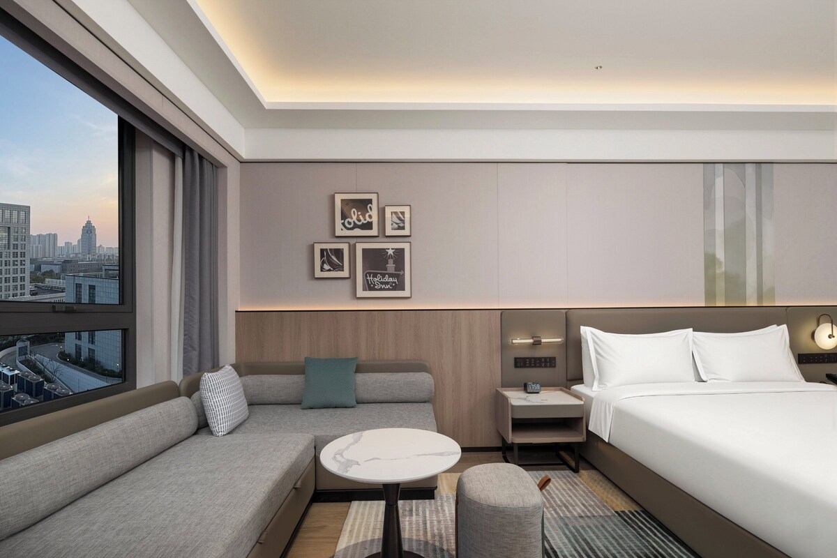 A modern hotel room with a sofa, a small round table, a bedside table with a clock, a bed, and a city view.