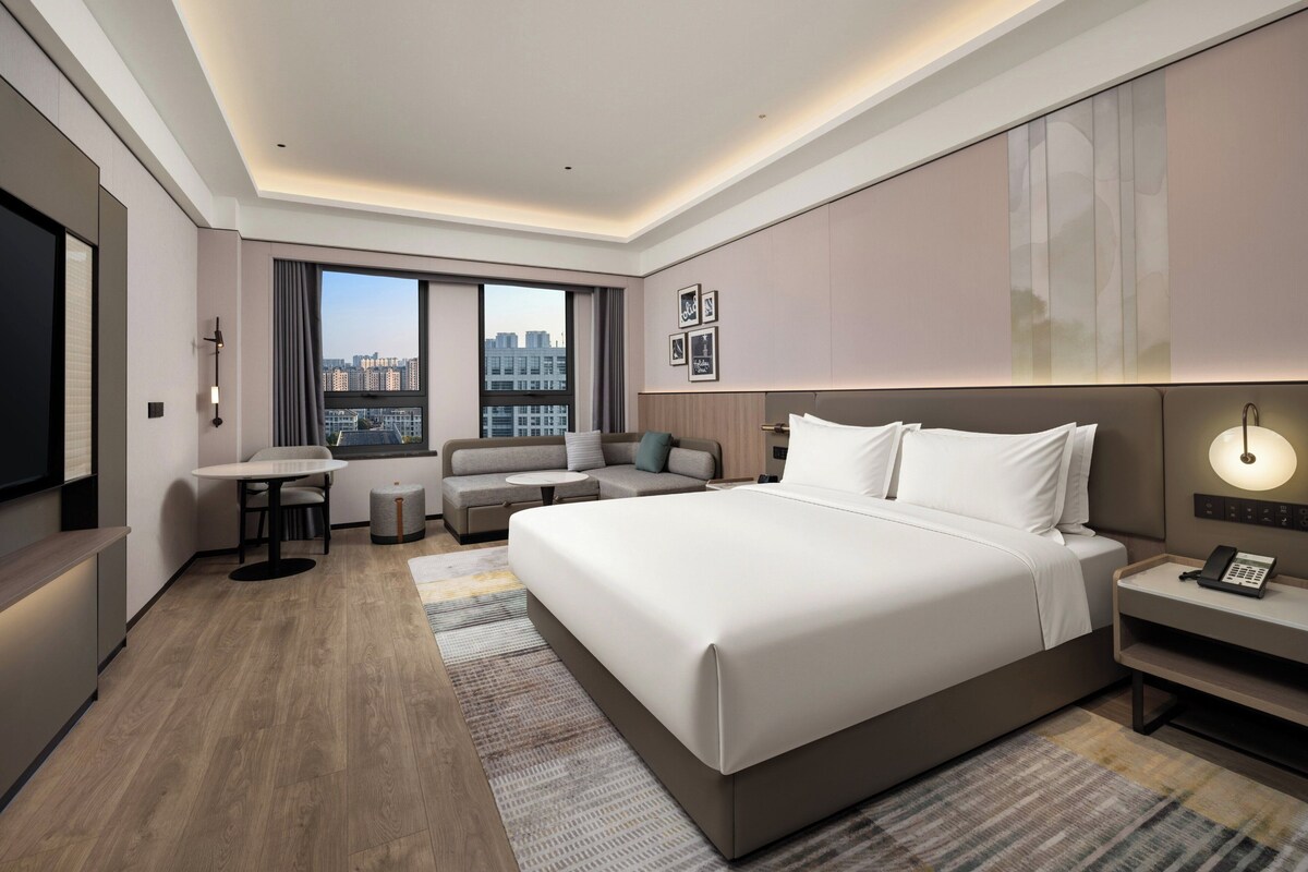 A modern hotel room with a large bed, a seating area, a desk, and a city view.