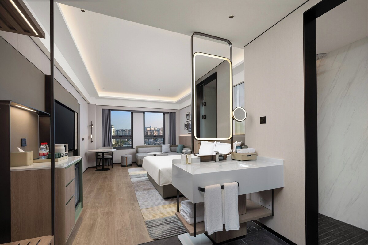 A modern hotel room with a large bed, a sitting area, a vanity with a mirror, and a view of the city.