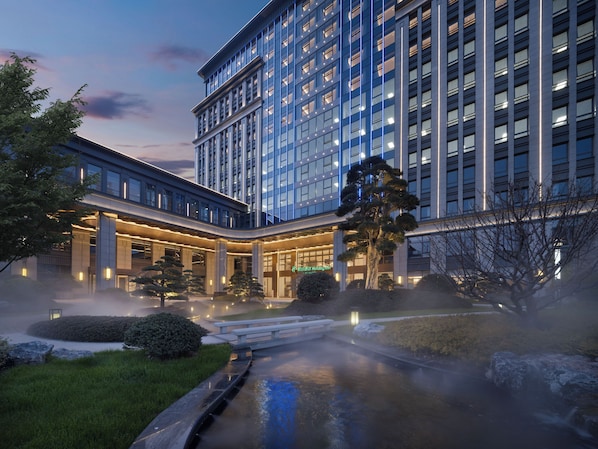 A modern hotel with a large glass facade, a courtyard with a water feature, and a well-lit entrance.