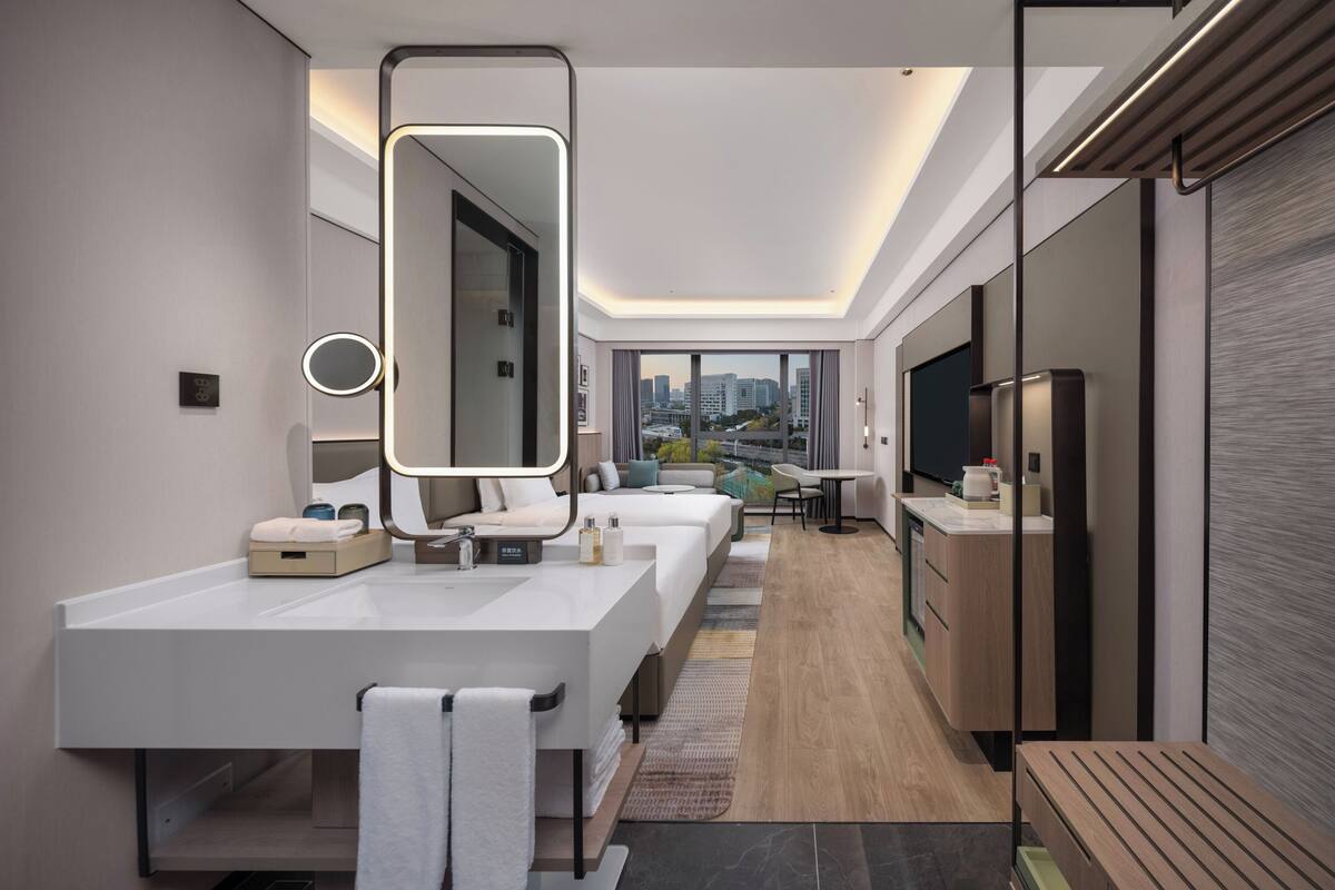 Un baño de hotel moderno con un espejo grande, dos lavamanos, un tocador y una zona de estar con vistas.