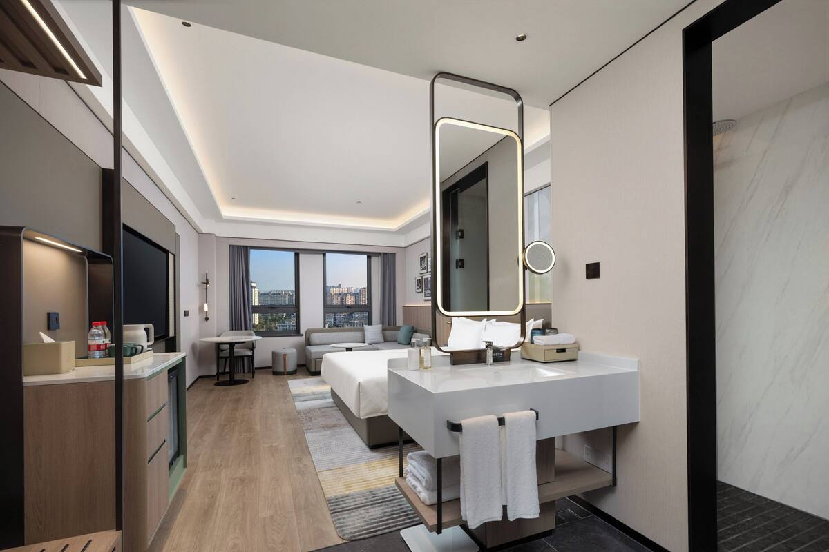 Una habitación de hotel moderna con una cama grande, una zona de estar, un tocador con espejo y vistas a la ciudad.