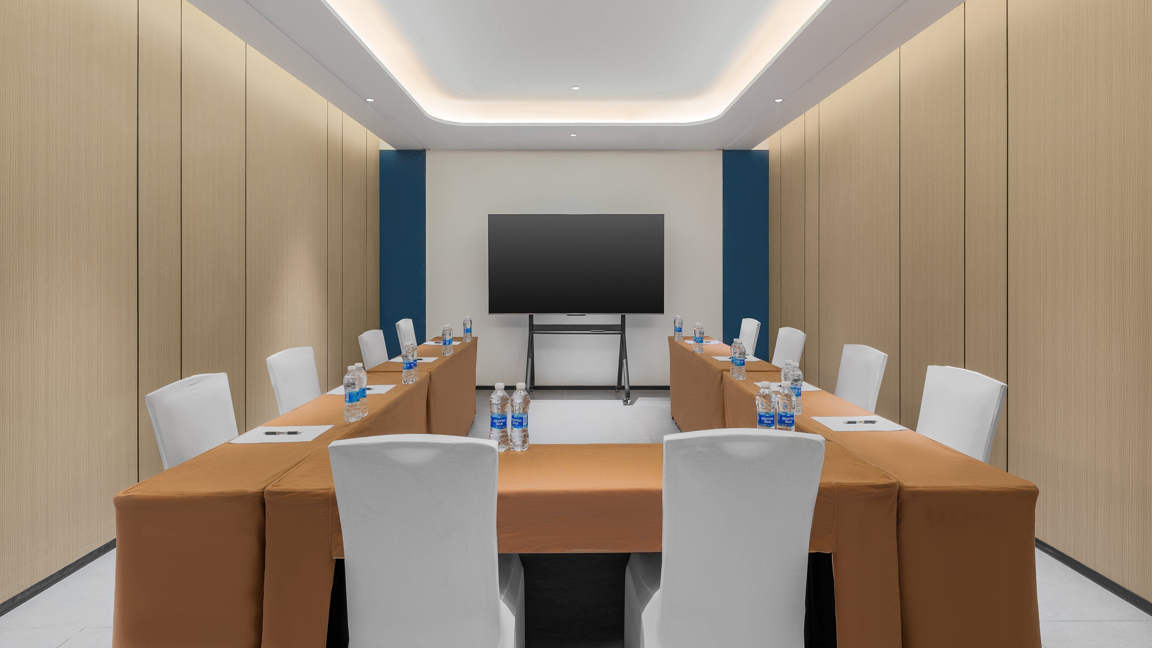Une salle de conférence avec une longue table, des chaises blanches, des bouteilles d’eau et un grand écran.