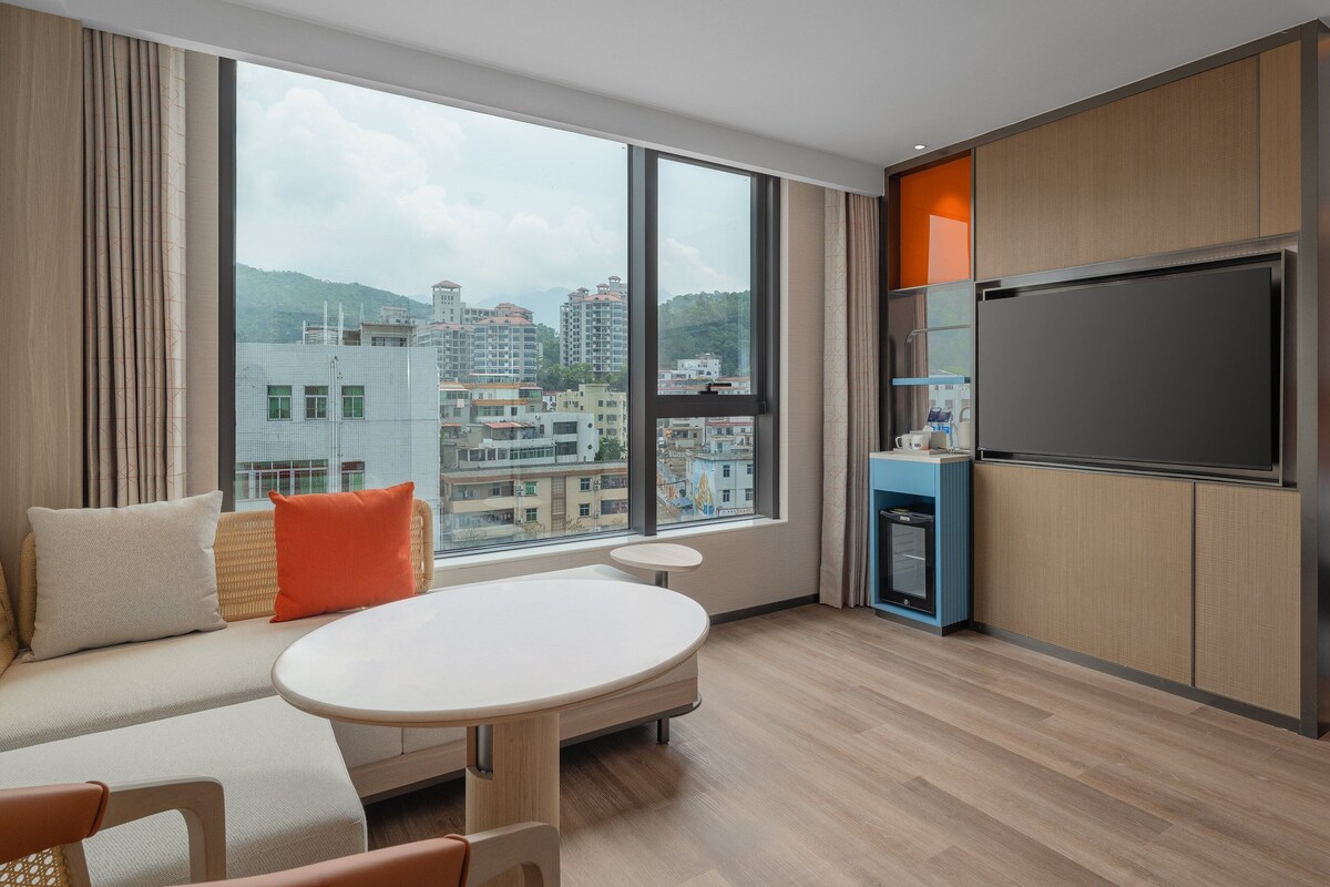 Habitación de hotel con un ventanal grande, cama, televisión, una mesita y zona de estar.