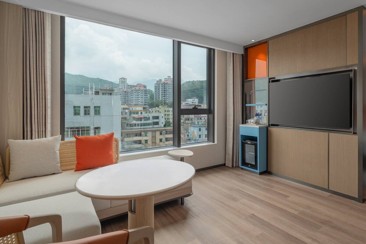 Une chambre d’hôtel avec une grande fenêtre, un lit, une télévision, une petite table et un coin salon.