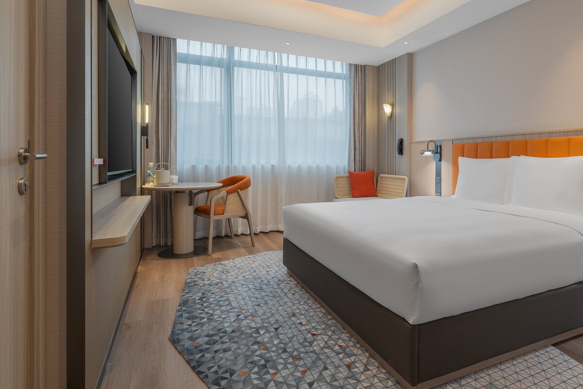 Una habitación de hotel con cama, escritorio, silla, ventana con cortinas y lámpara de pared.