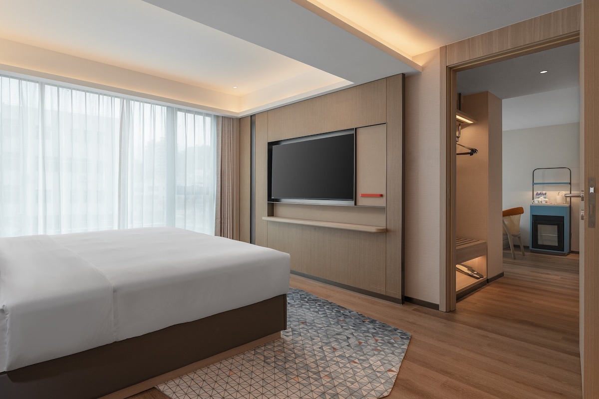 A modern hotel room with a large bed, a flat-screen TV, a bedside table, and a view of the city through the window.