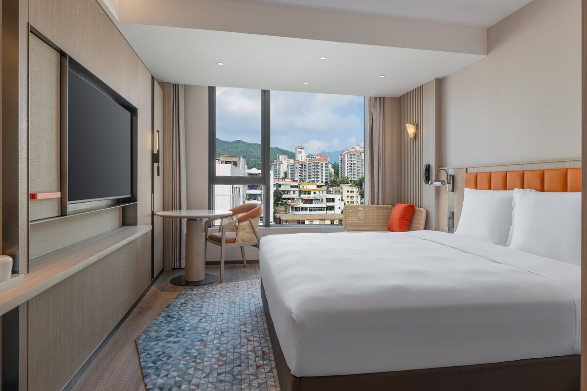Habitación de hotel con una cama grande, una mesita, una silla, un televisor y vistas a edificios.